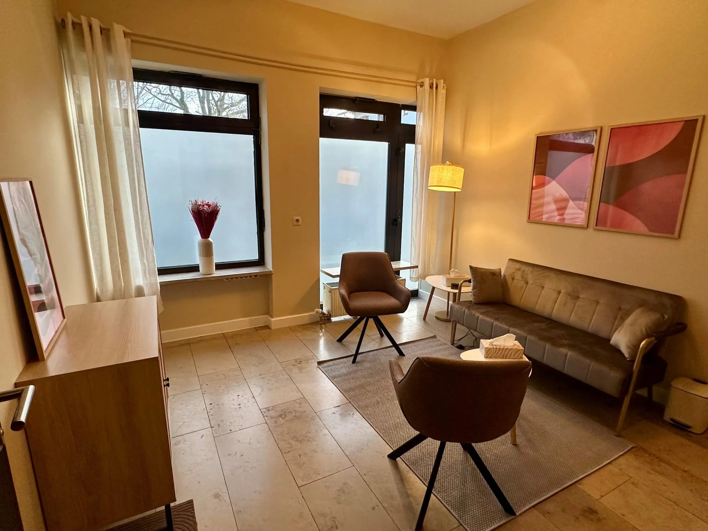 Gesprächsraum mit Sofa, zwei braunen Sesseln, Sideboard, Stehlampe, Teppich und zwei rosa Bildern an der Wand