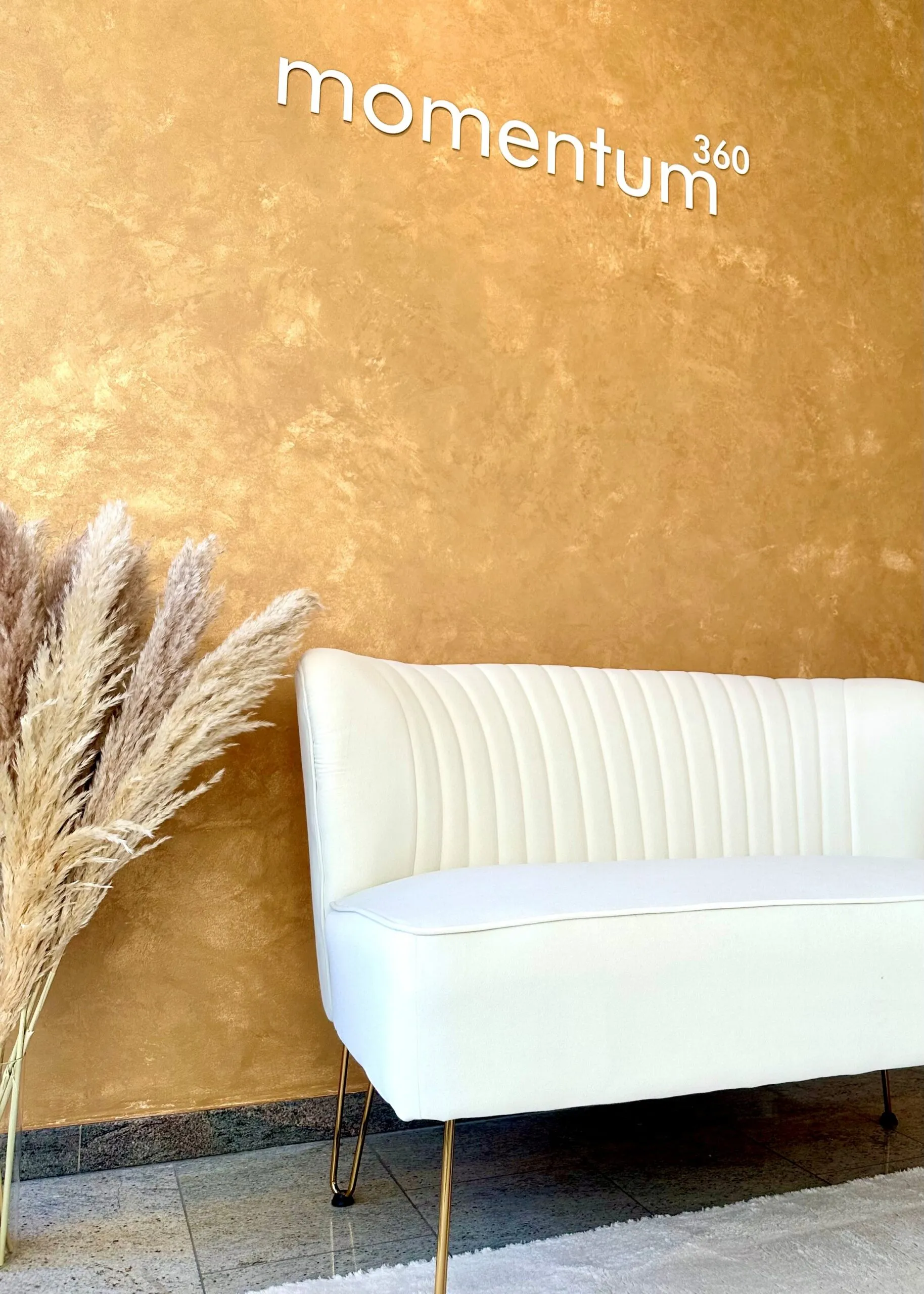 Goldene Wand mit momentum360-Schriftzug, davor ein weißes Sofa und Pampasgras